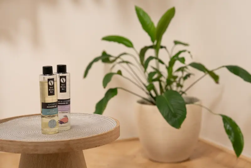 Two massage oil bottles on a wooden table with a green plant softly blurred behind them.